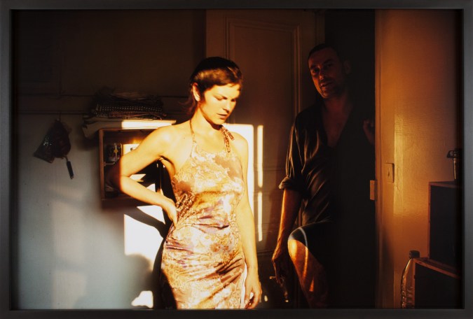 Color photograph of a young woman standing in sunlight from a window in front of a man in a shadowed doorway