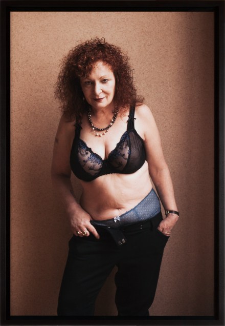 Color photograph of a woman in a lace bra and jeans with hands in her pockets standing in front of a beige wall