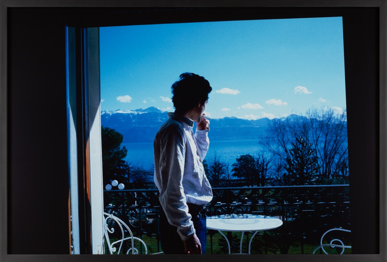 Color photograph of a figure smoking on a balcony and looking towards the horizon framed in black