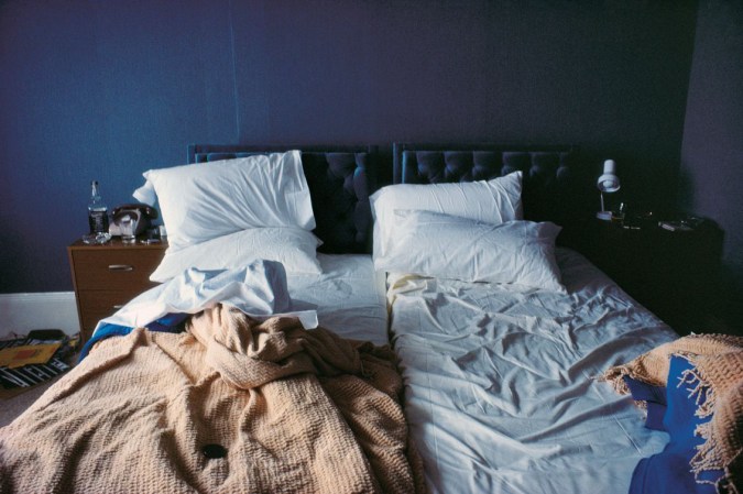 Color photograph of a bed with unmade sheets, against a blue wall
