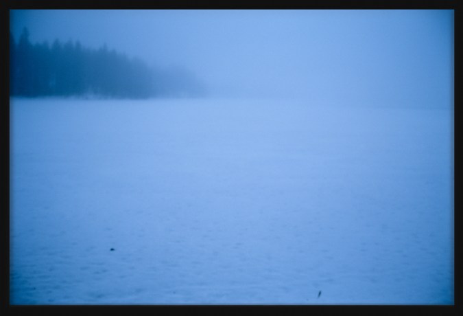 Color photograph of a snowy plain with trees on the horizon in a blue tint framed in black