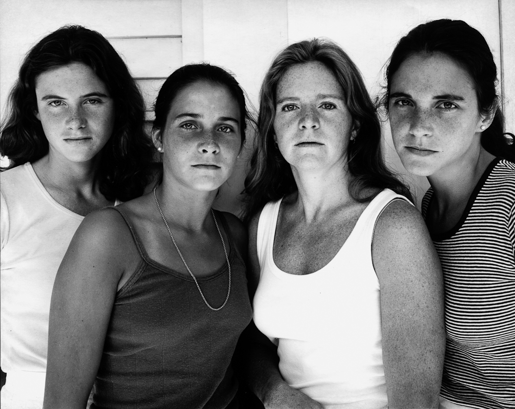 Black-and-white photographic portrait of four young women with long hair standing in front of a clapboard wall