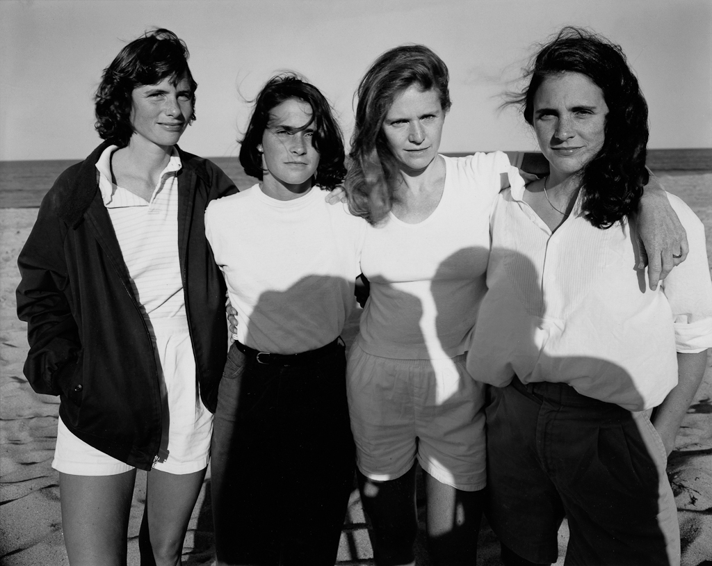 Black-and-white photographic portrait of four young women standing on a sunny beach with the ocean on the horizon