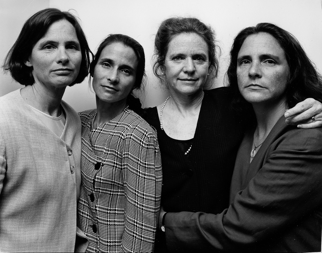 Black-and-white photographic portrait of four women with arms around each other standing in front of a blank wall