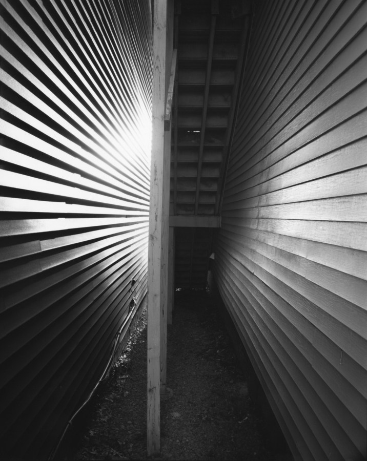 Black-and-white photograph of a stairway from beneath between two clapboard walls
