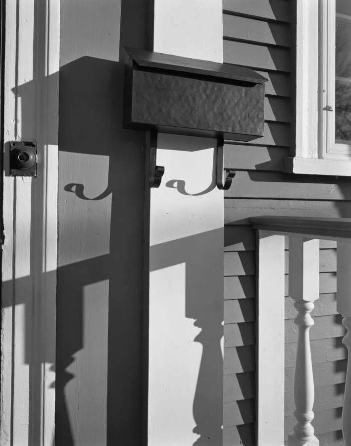 Black-and-white photograph of a metal mailbox on the wall of a clap-board sided house