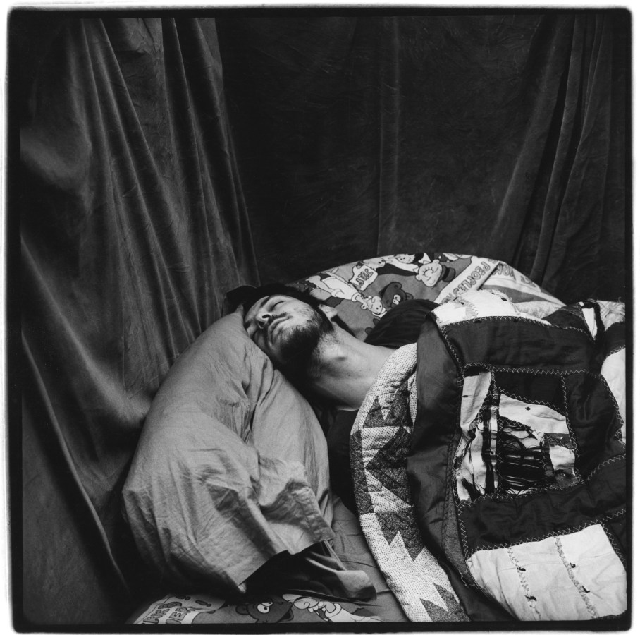 Black and white photograph of a male figure lounging in bed under sheets