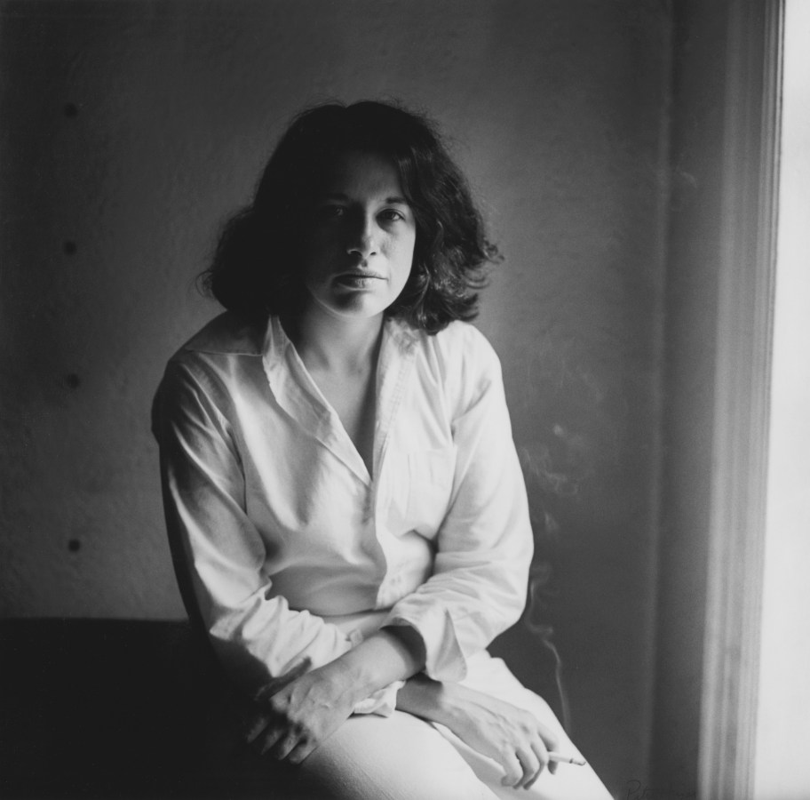 Black and white photograph of a seated figure in white button up, smoking a cigarette and looking at viewer