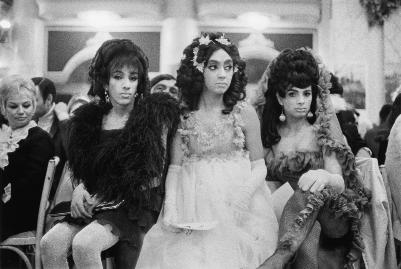 Black and white photograph of three seated figures in female drag in a ballroom