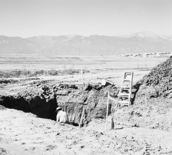A black and white photograph of a lone person digging with mountains in the background.