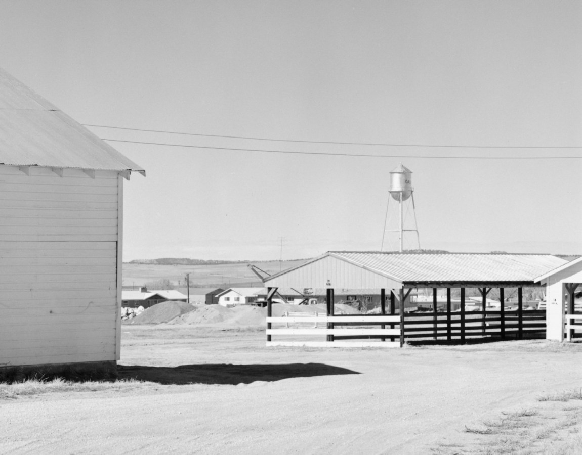 A black and white photograph of empty fairgrounds with a water tank in the distance and a brightly lit clear sky.