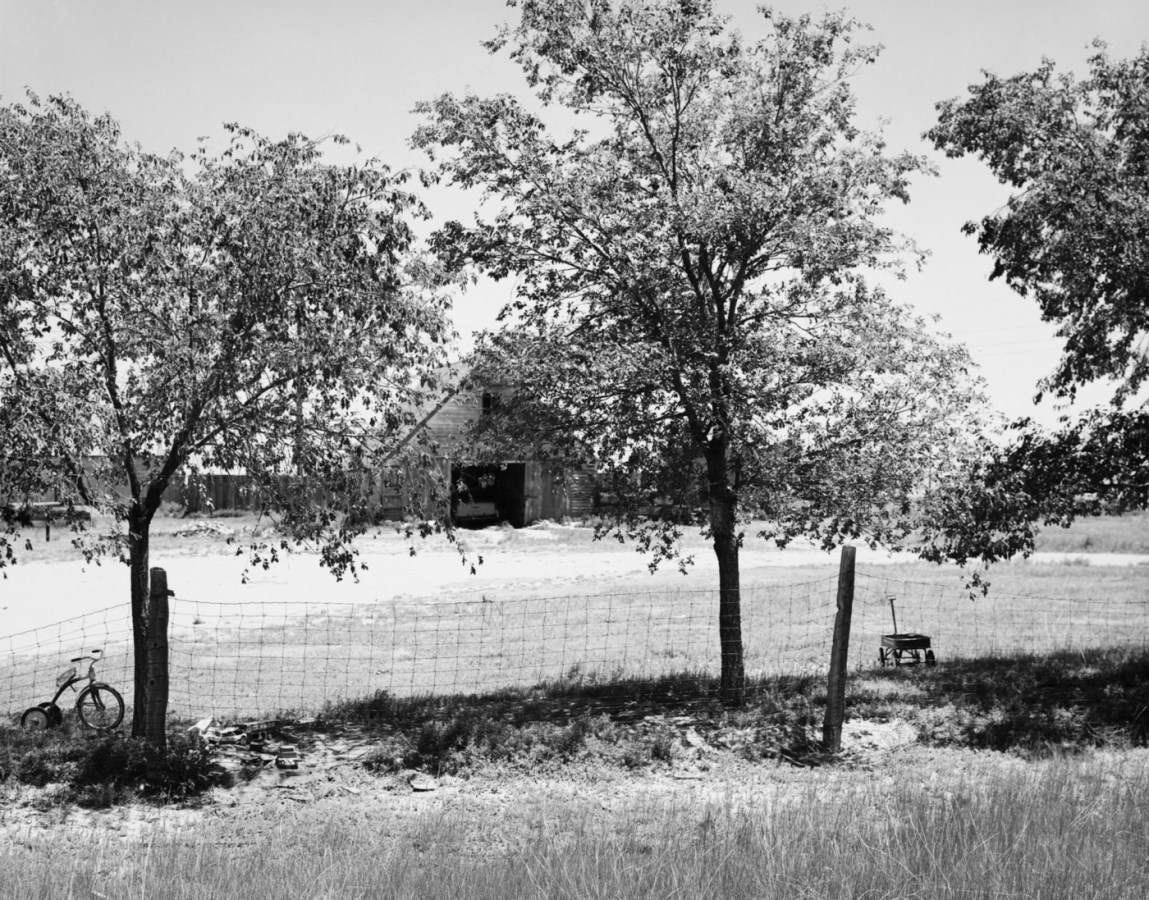 A black and white photograph with trees, a wire fence, a tricycle at the left side of the image, a wagon at the right side of the image and a barn in the background.