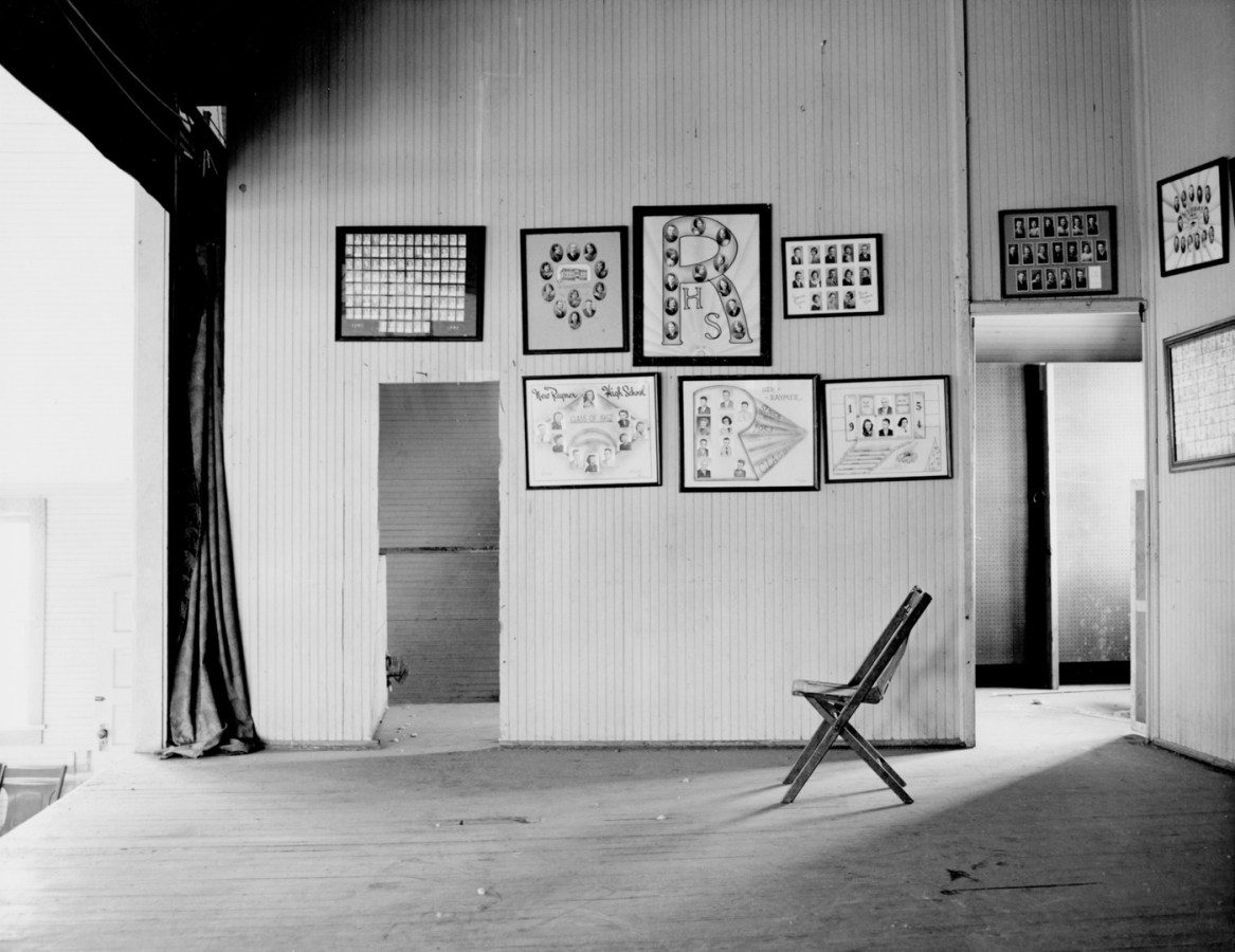 An empty stage with multiple framed photographs of graduating students, a curtain at the left side of the image and single wooden folding chair at right side of the image.