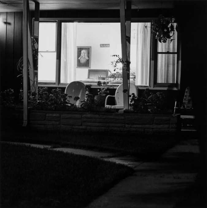 Black and white photograph of the front of a suburban home at night, with two chairs on the front porch and the home interior visible through the window.