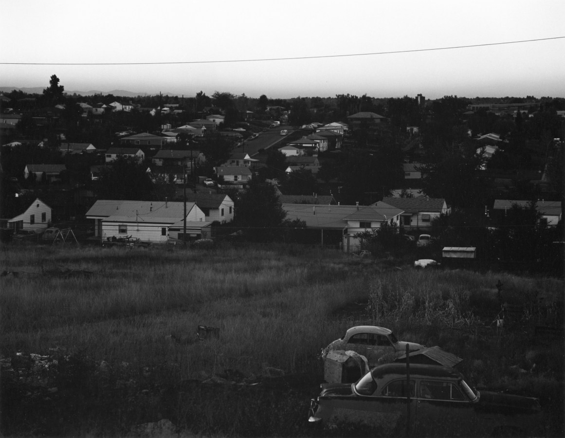 Black and white photograph of a suburban neighborhood adjacent to a field with a few abandoned cars