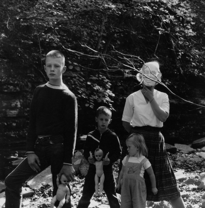 Black-and-white photograph of three children holding baby dolls and a woman holding a mask over her face