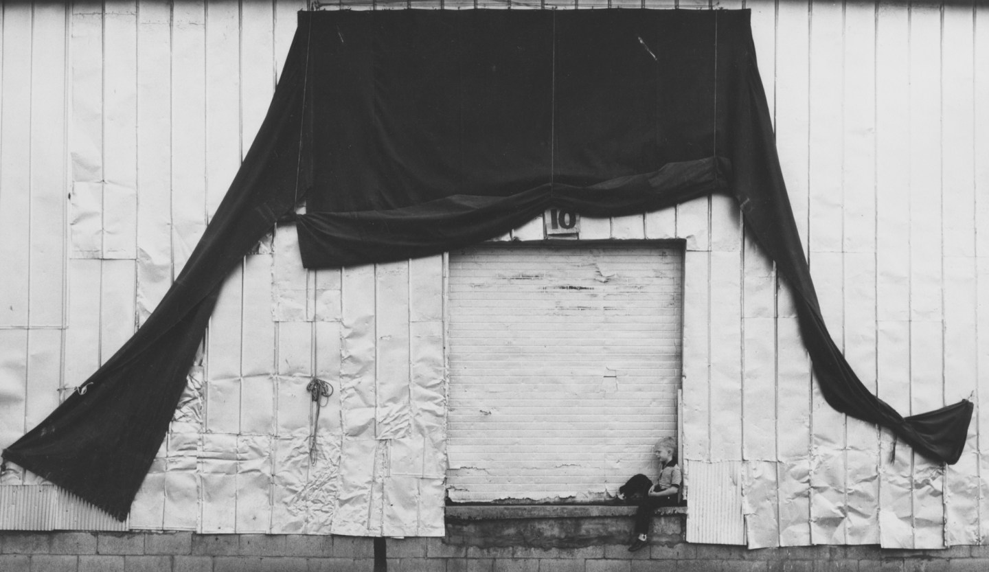 Black-and-white photograph of a young boy sitting in front of a shuttered loading dock