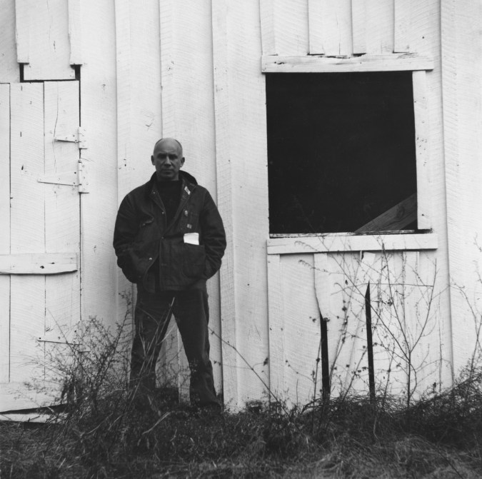 Black and white photograph of a male figure standing in front of white wooden structure