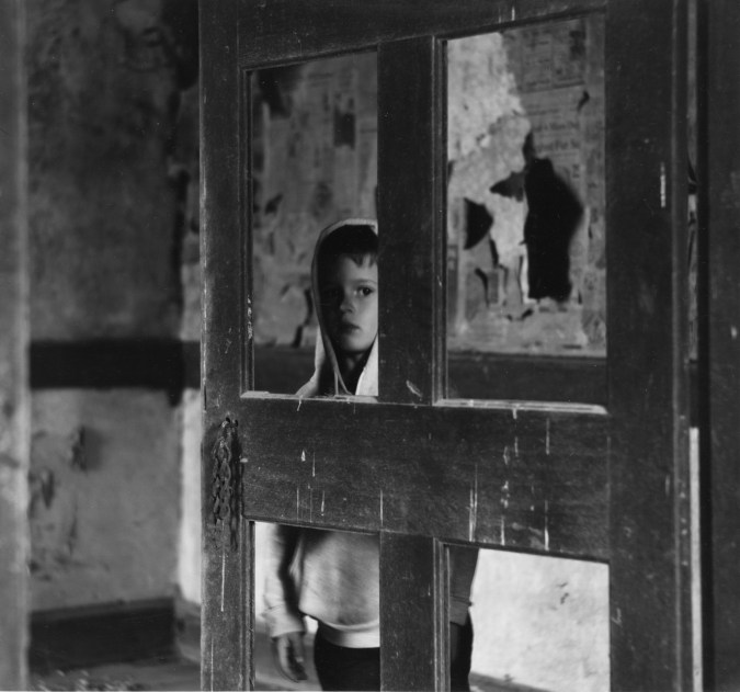 Black-and-white photograph of a young boy in a hooded sweatshirt looking through a door with missing panels from a decrepit room