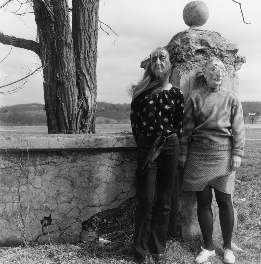 Black and white photograph of two female figures with masks standing by concrete column outdoors