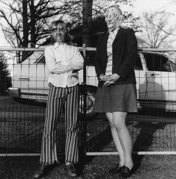 Black-and-white photograph of two people in rubber masks standing in front of a wire fence