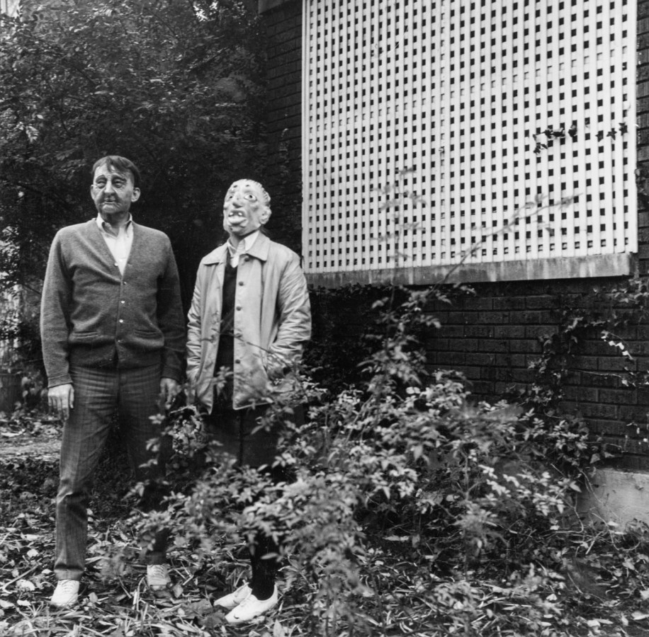 Black-and-white photograph of two people in rubber masks standing next to a lattice-in porch of a brick building
