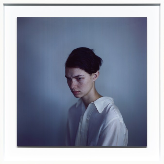 Color photographic portrait of a young woman with tied up dark hair and a somber downcast expression