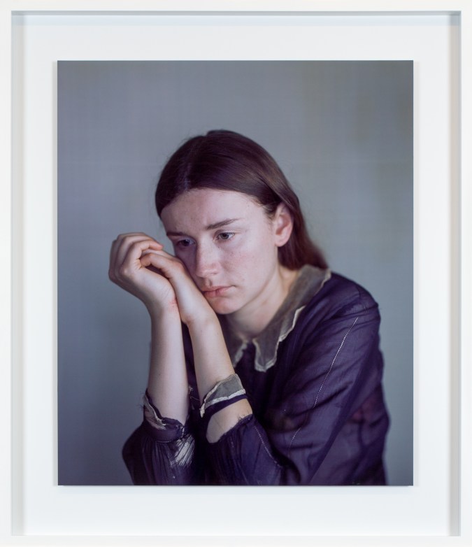 Color photographic portrait of a young woman in a purple shirt with her cheek resting on her clasped hands