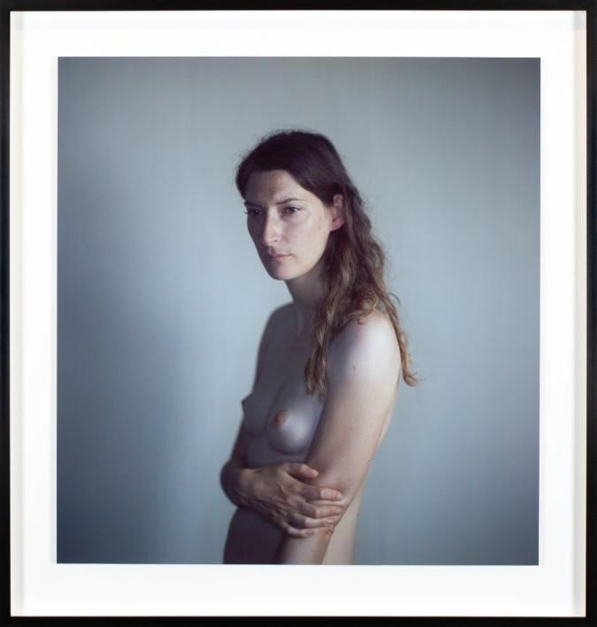 Color photographic portrait of a topless young woman against a gray background