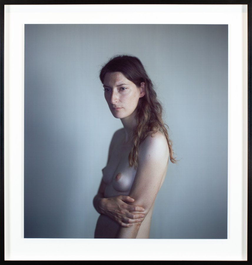 Color photographic portrait of a topless young woman against a gray background