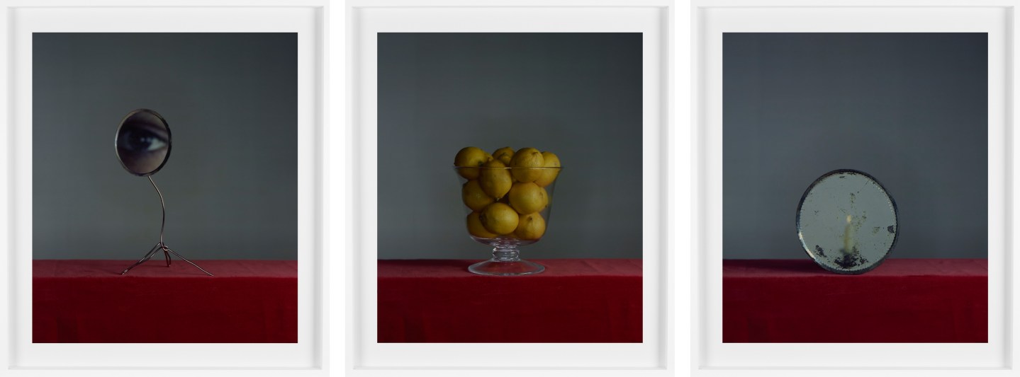 Triptych of color still life photographs depicting a small circular mirror with eye, a glass bowl of lemons, and a small circular mirror with candle all in white frames