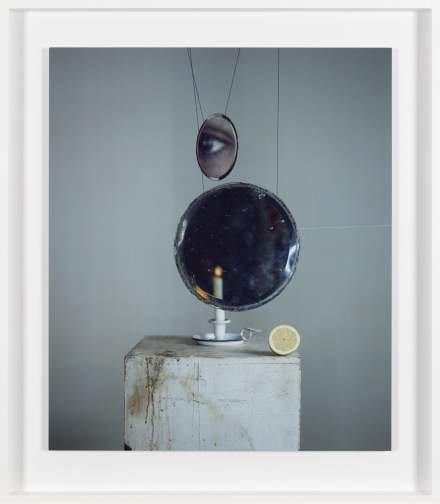 Color photograph of a candle and lemon on plinth with suspended glass and mirror reflecting an eye framed in white