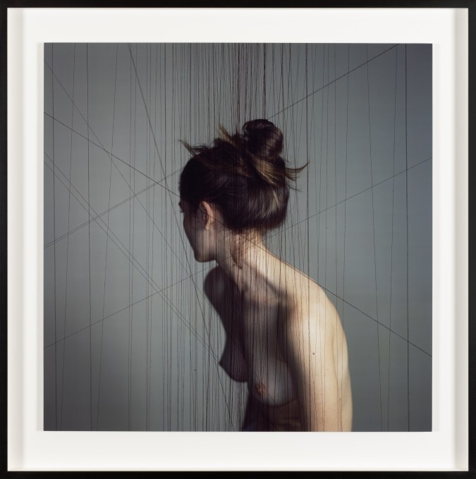 Color photograph of a nude women turned away from camera amongst suspended black thread in black frame