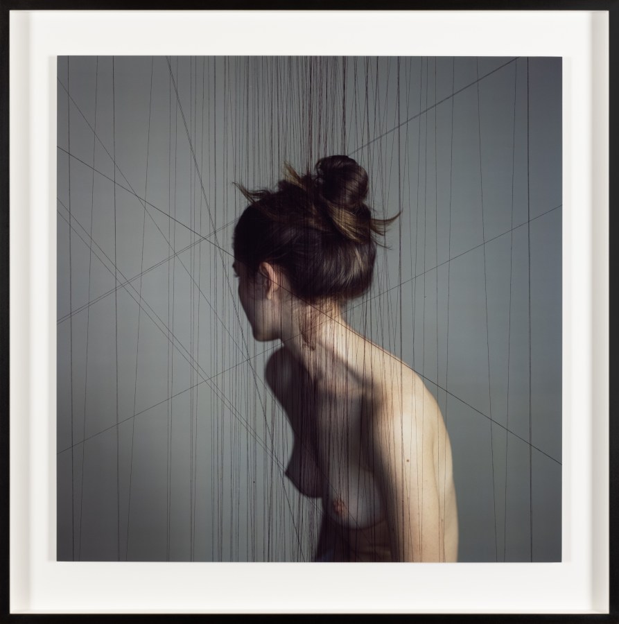 Color photograph of a nude women turned away from camera amongst suspended black thread in black frame