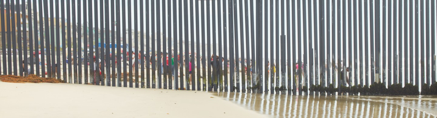 Color photograph of a tall metal fence running into the ocean from the beach, with beach goers visible through the bars