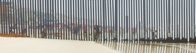 Color photograph of a tall metal fence running into the ocean from the beach, with beach goers visible through the bars