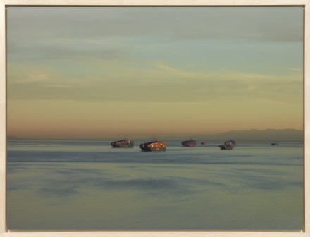 Color photograph framed in bleached wood depicting several cargo ships on the water close to sunset