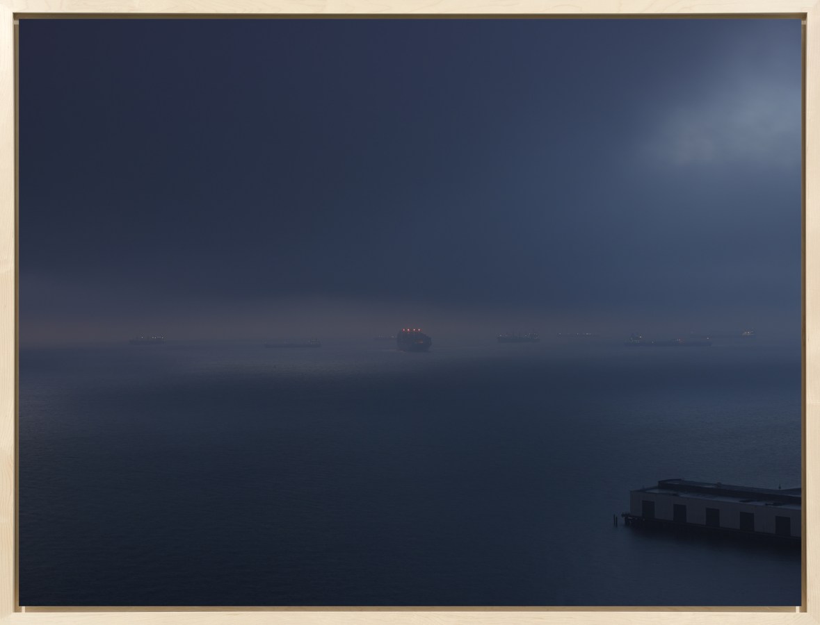 Color photograph of cargo ships moving across the water at around sunrise on an overcast morning framed in a bleached wood frame