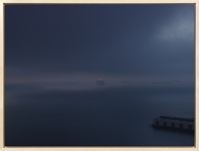 Color photograph of cargo ships moving across the water at around sunrise on an overcast morning framed in a bleached wood frame