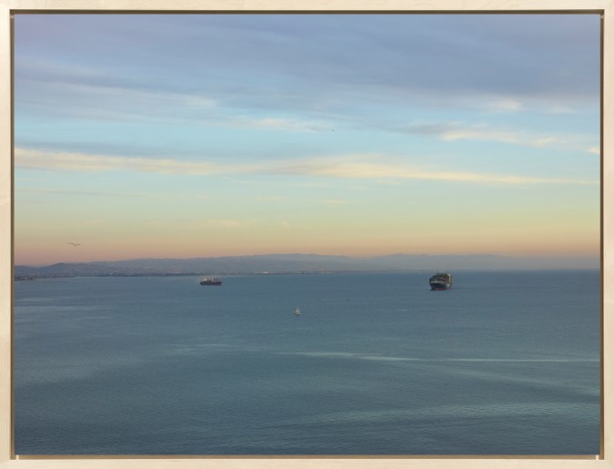Color image of a color photograph of two cargo ships on the water with birds and a sailboat framed in bleached wood frame