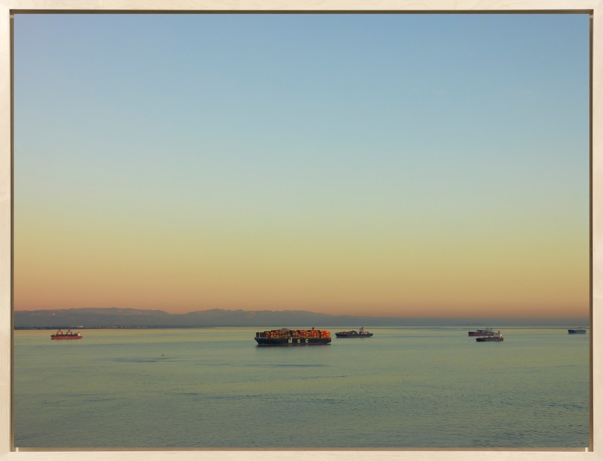 Color image of a color photograph of six cargo ships traveling along the water on a clear day framed in bleached wood