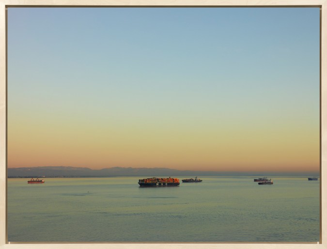 Color image of a color photograph of six cargo ships traveling along the water on a clear day framed in bleached wood