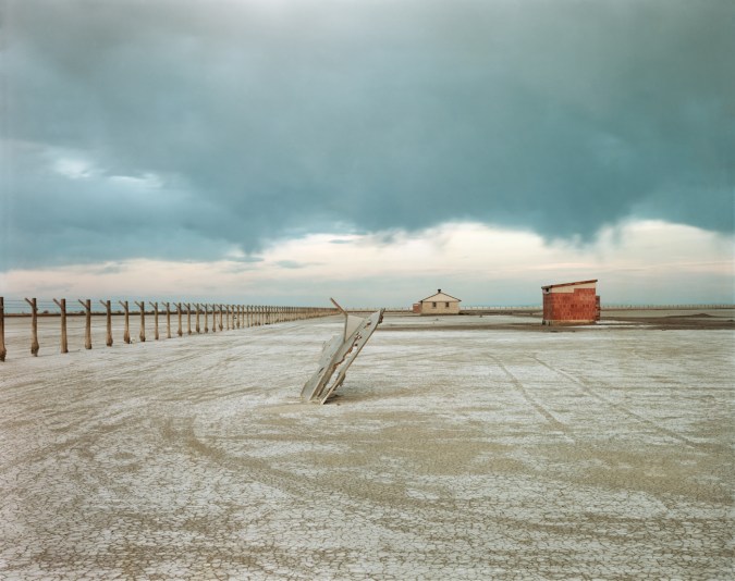 Color photograph of a rusted piece of metal sticking out of the ground at an angle in a bare field with two small buildings in the background