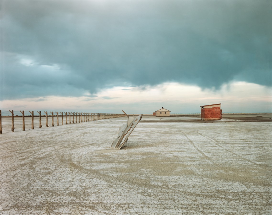 Color photograph of a rusted piece of metal sticking out of the ground at an angle in a bare field with two small buildings in the background