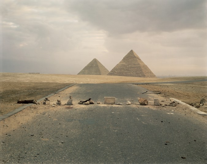 Color photograph of scattered rocks and blocks across a road leading towards two pyramids on the horizon