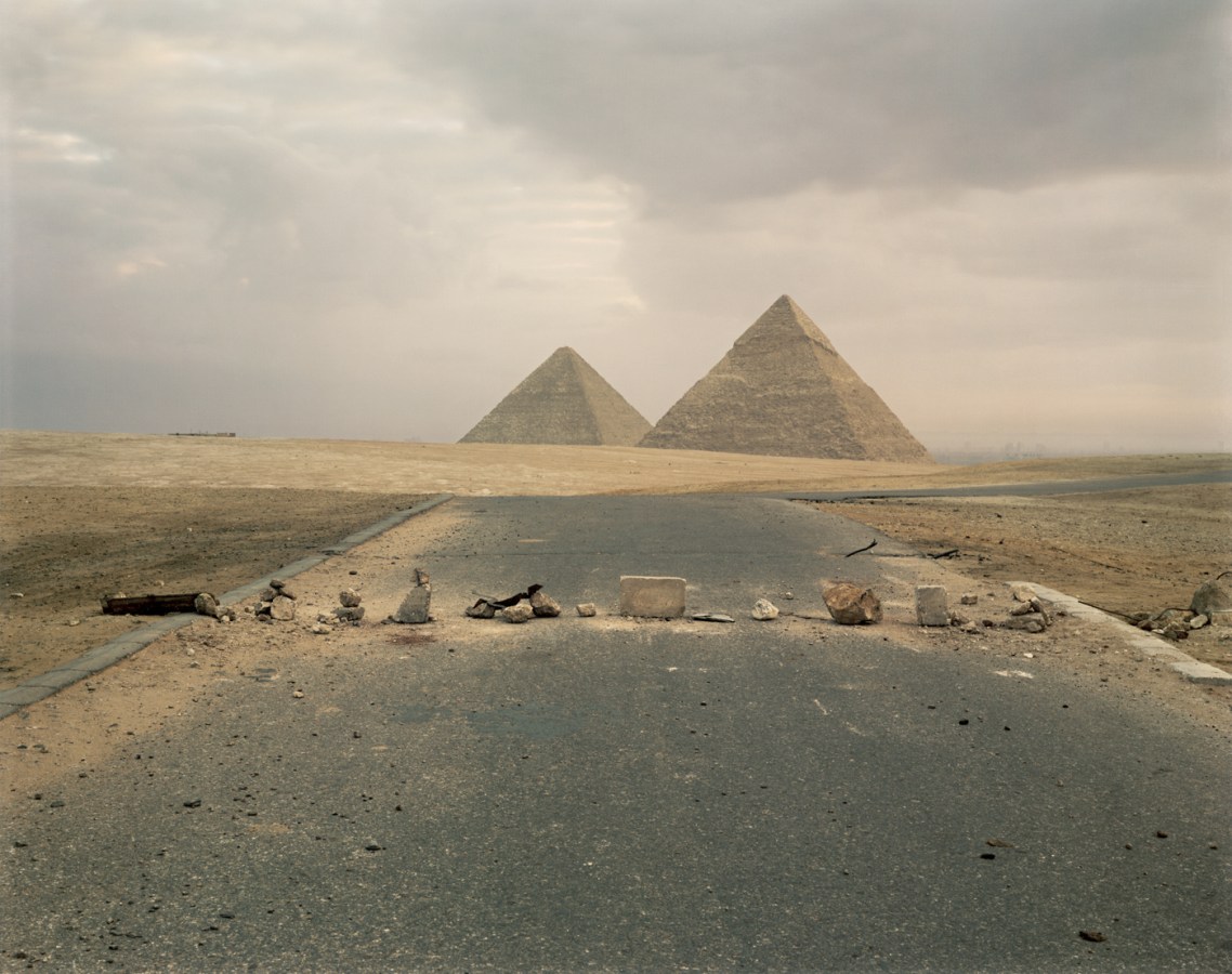 Color photograph of scattered rocks and blocks across a road leading towards two pyramids on the horizon
