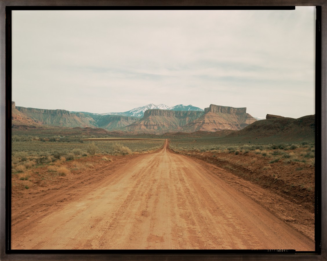 Color photograph of a road receding into the distant desert with red-orange tablelands and snow-capped mountains on the horizon