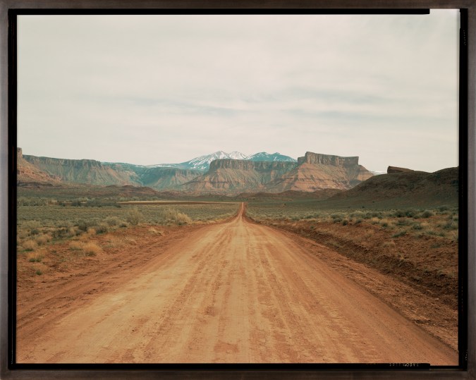 Color photograph of a road receding into the distant desert with red-orange tablelands and snow-capped mountains on the horizon
