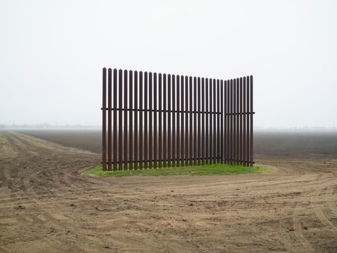 Color photograph of a segment of tall metal fence in the middle of a bare field