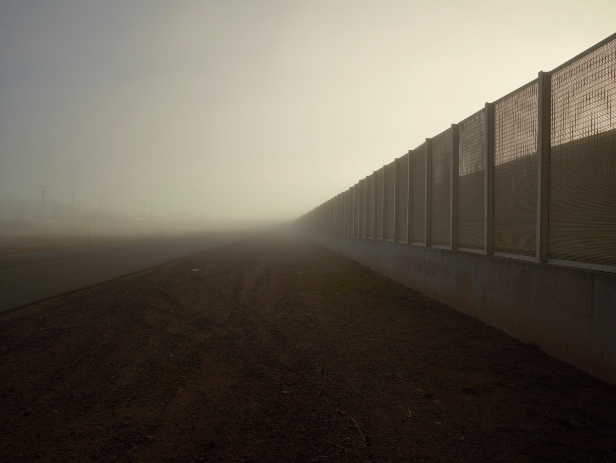 Color photograph of a tall metal and concrete fence receding into the misty horizon on the right